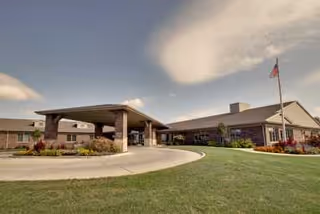 Exterior view of a single-story senior living facility building with a covered entrance driveway, well-maintained lawn, flower beds, and an American flag on a flagpole under a partly cloudy sky.