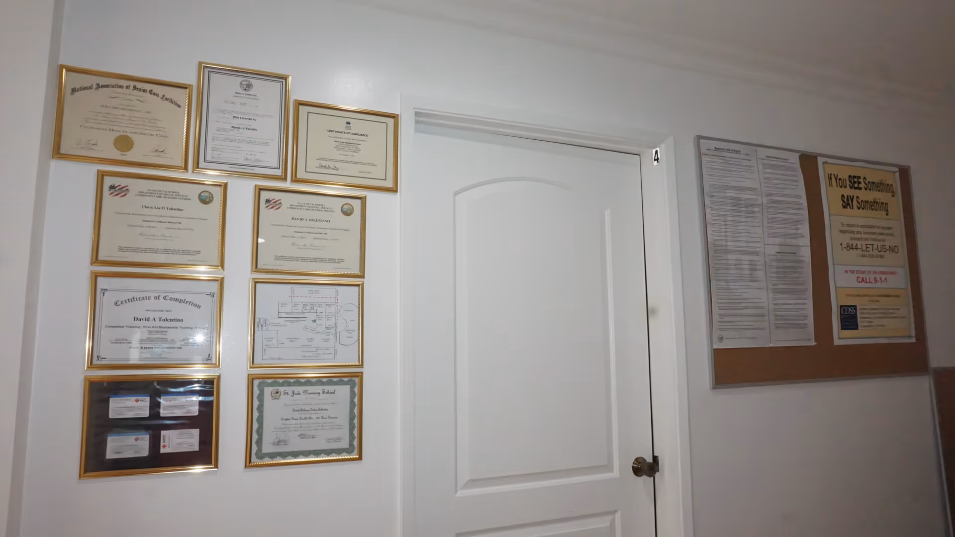 Interior hallway wall of Olive Branch Assisted Living with a white door labeled number 4. The wall features multiple framed certificates and documents on the left side and a bulletin board with notices on the right side.