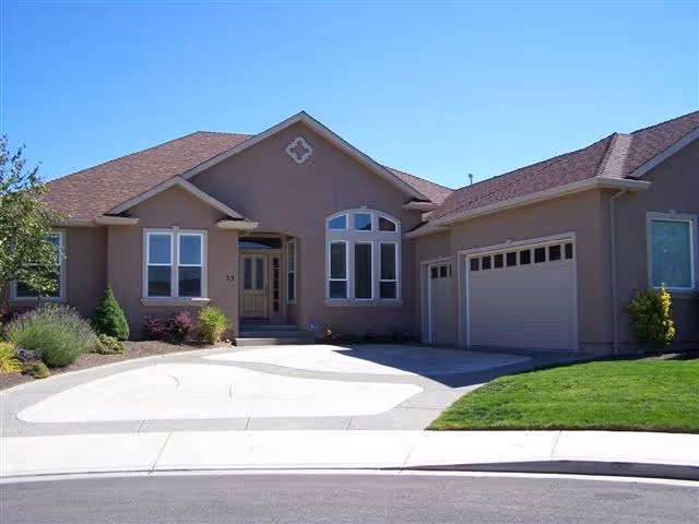 Front exterior view of a single-story house with a brown roof and beige walls, featuring a driveway, garage doors, and a small landscaped garden with shrubs and grass.