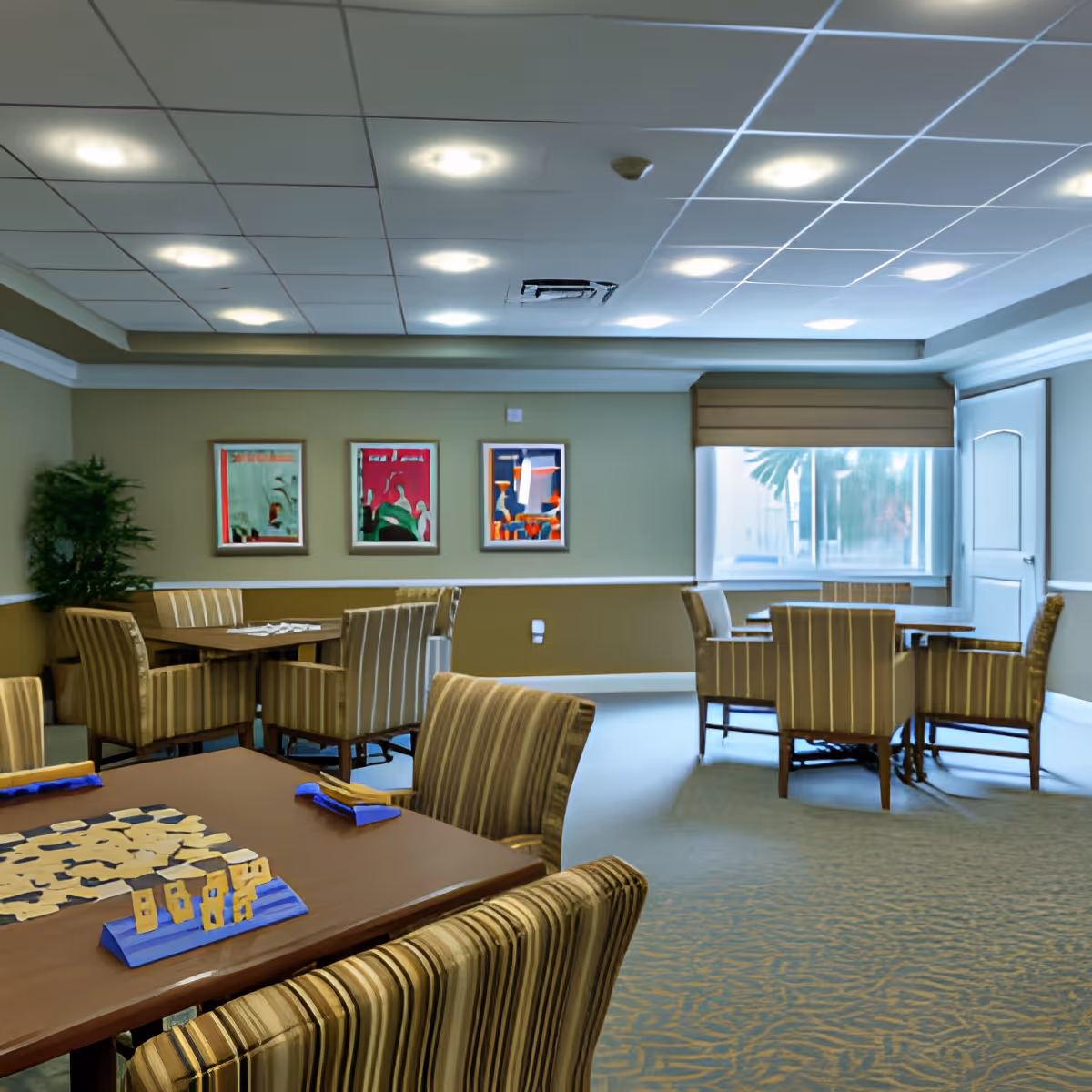 A well-lit room with multiple tables and striped upholstered chairs arranged for group seating. The room has a beige and light green color scheme with three colorful framed artworks on the wall and a large window with a beige blind partially drawn. A board game is set up on the closest table.