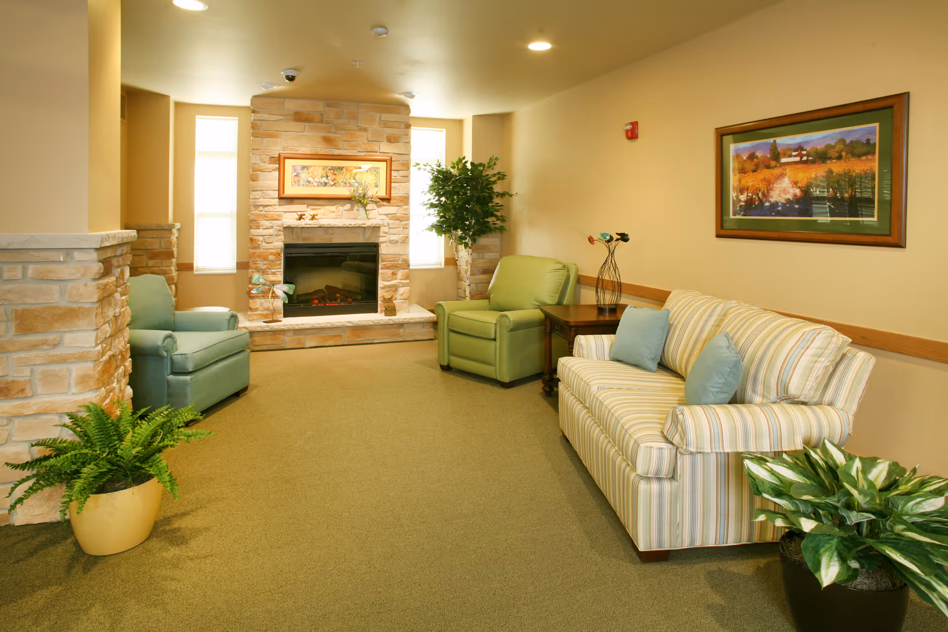 A cozy living room area with a stone fireplace in the center, flanked by two narrow windows. The room features a striped sofa with two blue pillows, a green armchair, and a light blue armchair. There are two potted plants on the floor and a framed landscape painting on the wall above the sofa. The walls are painted beige, and the carpet is green.