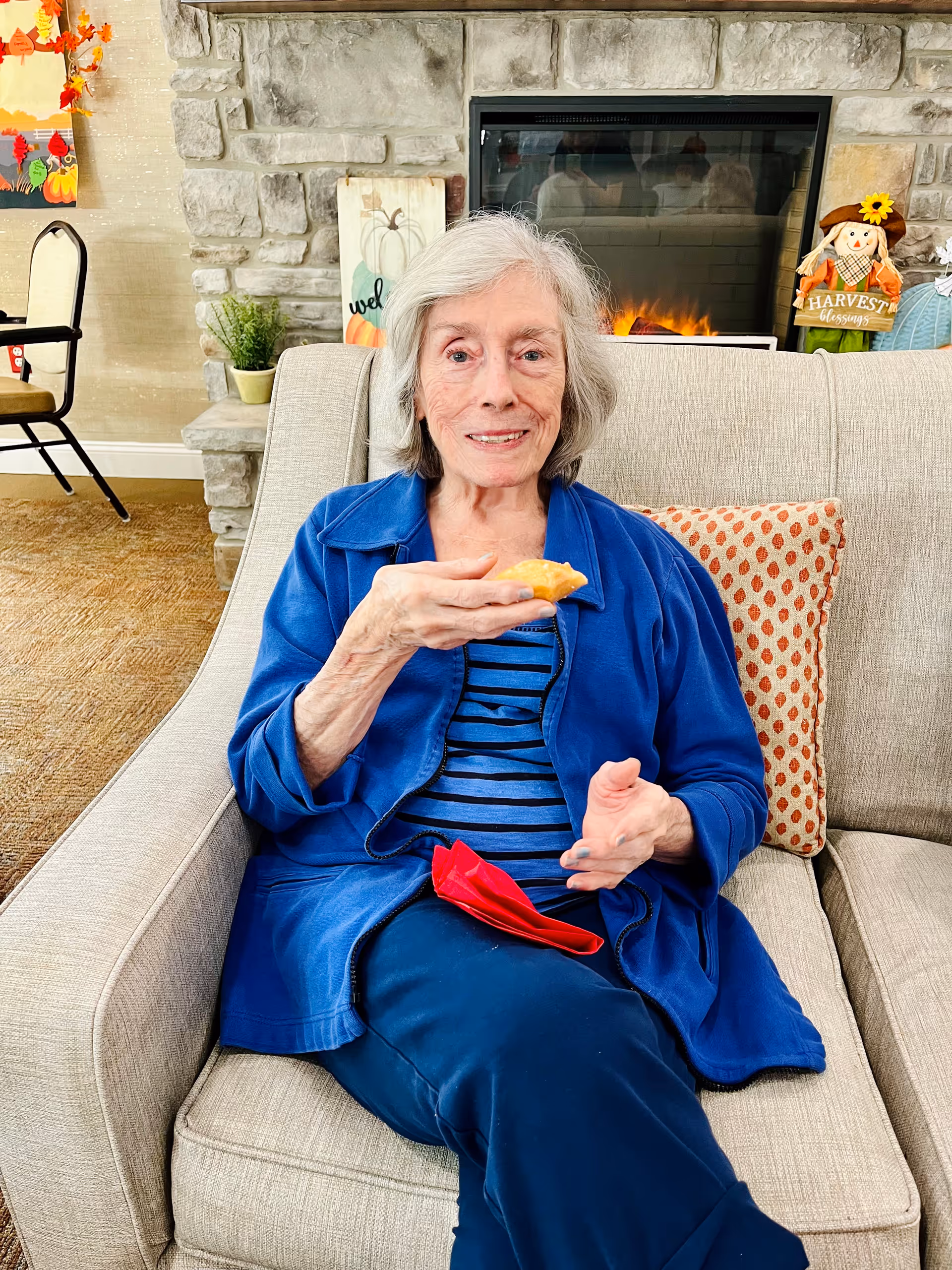 An elderly woman with gray hair wearing a blue jacket and striped shirt is sitting on a beige couch holding a piece of food. Behind her is a stone fireplace with a fire burning, a decorative sign with pumpkins, and a scarecrow decoration with a 'Harvest Blessings' sign. There is a patterned pillow on the couch and a chair and fall-themed decorations in the background.