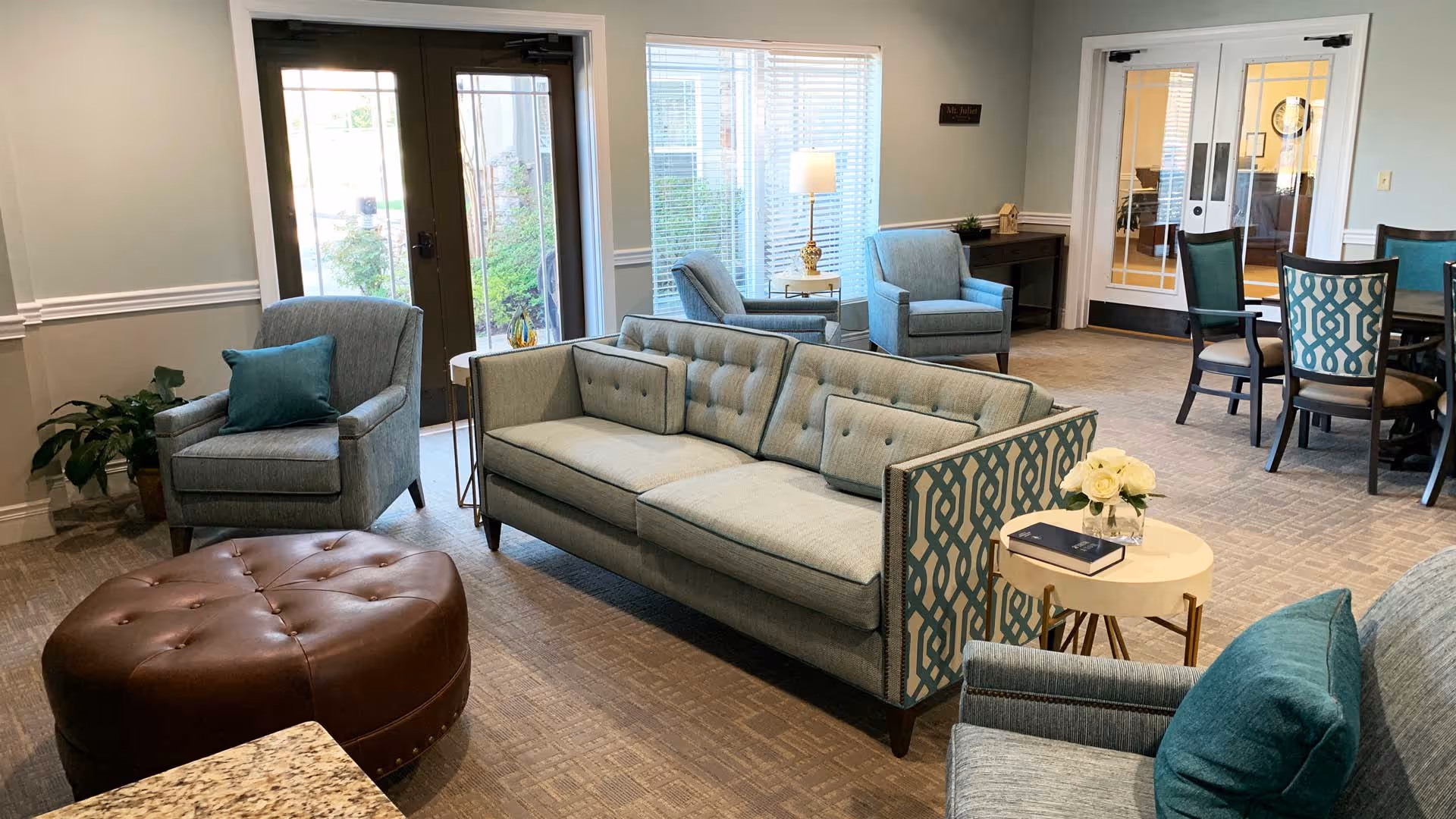 A bright and cozy living room area in a senior living facility featuring a light blue sofa with patterned sides, matching armchairs with teal pillows, a round brown leather ottoman, a small white side table with a vase of white flowers and a book, and a dining table with chairs in the background. Large windows and glass doors allow natural light to fill the space.