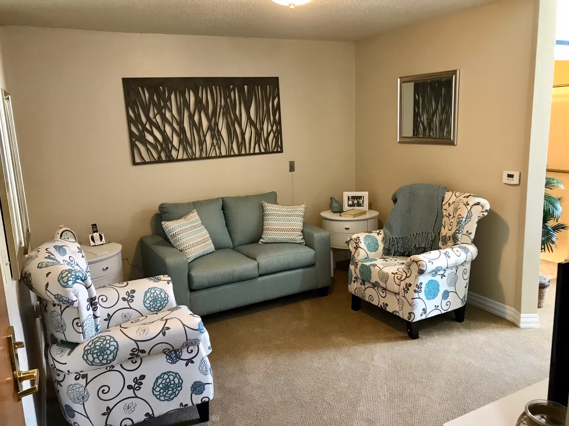 Small living room with a teal loveseat, two patterned armchairs, side tables, and wall decor.