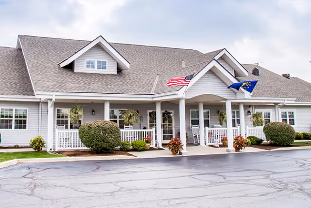 Front exterior view of a single-story building with a gray roof and white siding, featuring a covered porch with white railings, hanging plants, and two flags—an American flag and a state flag—mounted near the entrance. The building is surrounded by neatly trimmed bushes and a paved driveway in front.