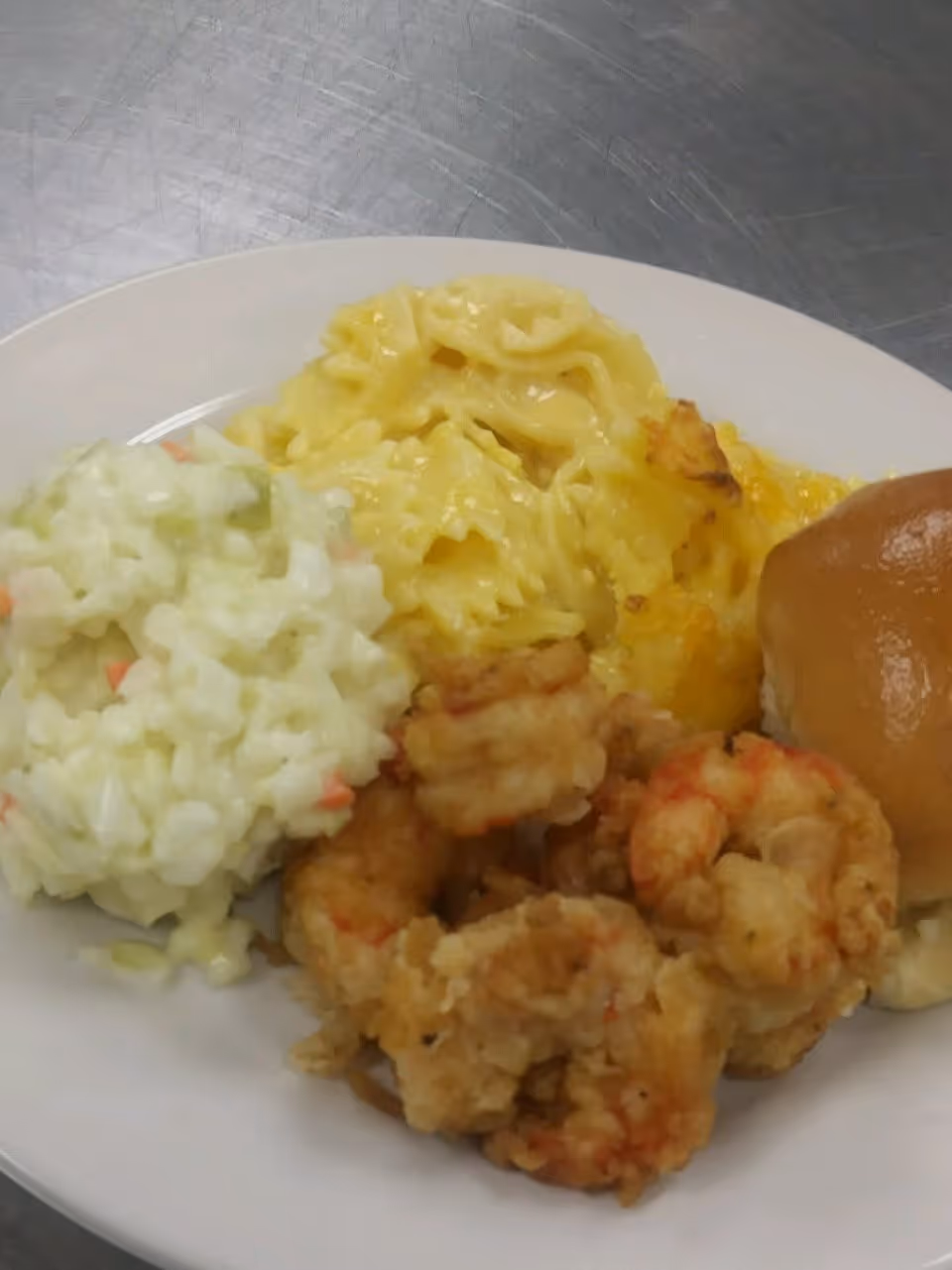 A white plate with a serving of fried shrimp, macaroni and cheese, a bread roll, and a side of creamy coleslaw on a metal surface.