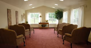 A bright and spacious living room with large windows letting in natural light. The room features six upholstered armchairs arranged in two rows facing each other, a small wooden table with a lamp and decorative plants near the windows, and a large potted plant in the corner. The carpet is a deep red color, and the walls are light-colored with three framed artworks.