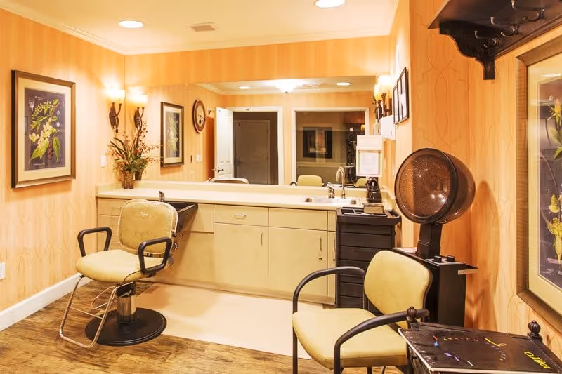 Small salon-style interior with two styling chairs, a countertop with sink and mirror, and a hooded hair dryer.