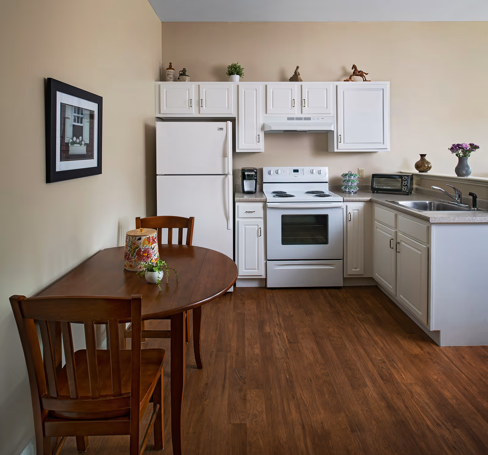 A small kitchen area with white cabinets, a white refrigerator, a white electric stove with oven, a coffee maker, a toaster oven, and a double sink. There is a wooden dining table with two chairs, a decorative container, and a small plant on the table. The walls are beige, and there is a framed picture hanging on the wall. The floor is wood.