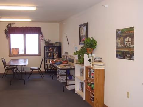 A communal interior room with tables and chairs, bookshelves stocked with books and games, potted plants, and wall art.