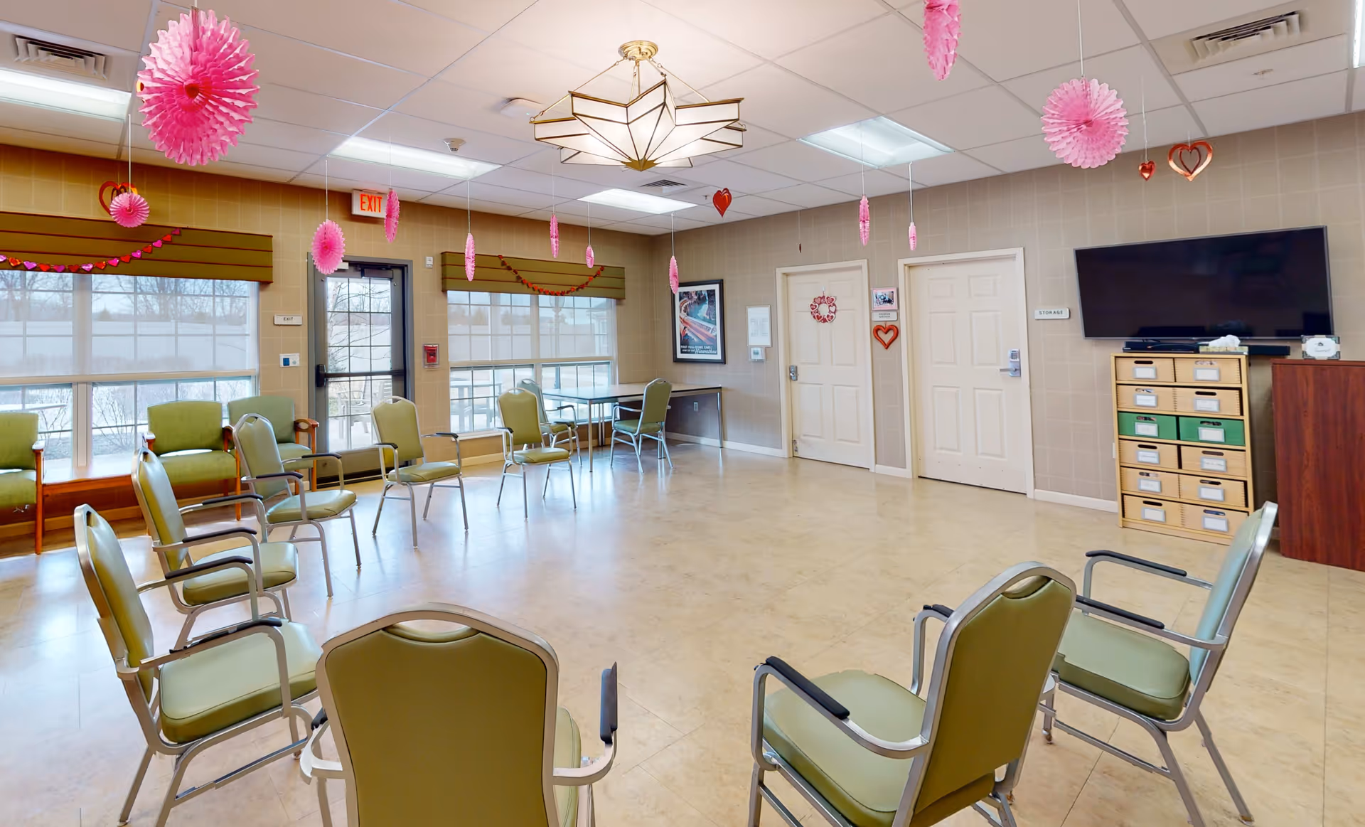A spacious room with chairs arranged in a circle, decorated with pink hanging paper decorations and heart-shaped ornaments. The room has large windows with blinds, a TV mounted on the wall, and a cabinet with labeled drawers. There are two closed doors and a table with chairs near the windows.