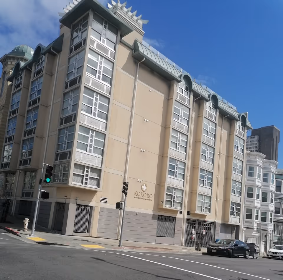 Exterior view of a multi-story building named Kokoro Assisted Living Inc, located at a street corner with traffic lights and parked cars under a clear blue sky.