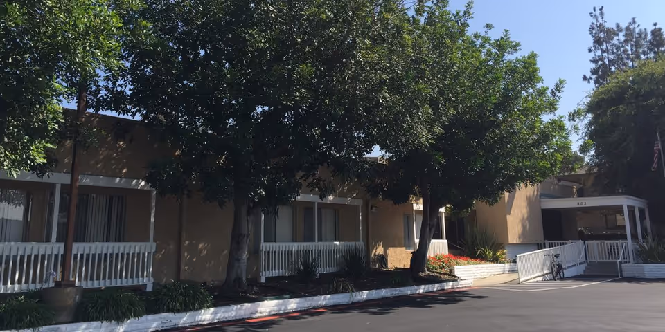 Exterior view of Monte Vista Healthcare Center showing a single-story building with beige walls, white railings, and several windows partially shaded by large green trees. There is a paved parking area in front with a bicycle parked near the entrance ramp.