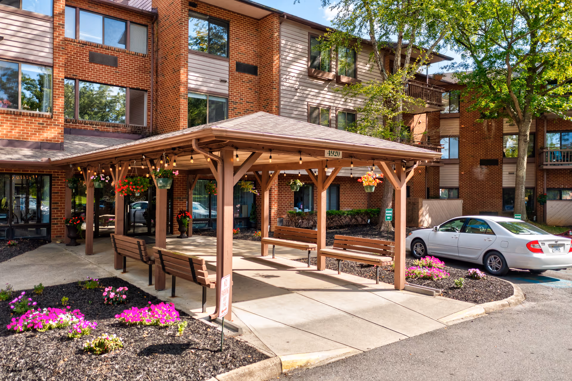 Covered wooden entry pavilion with benches and hanging flower baskets in front of a multi-story brick senior living building and landscaped flower beds.