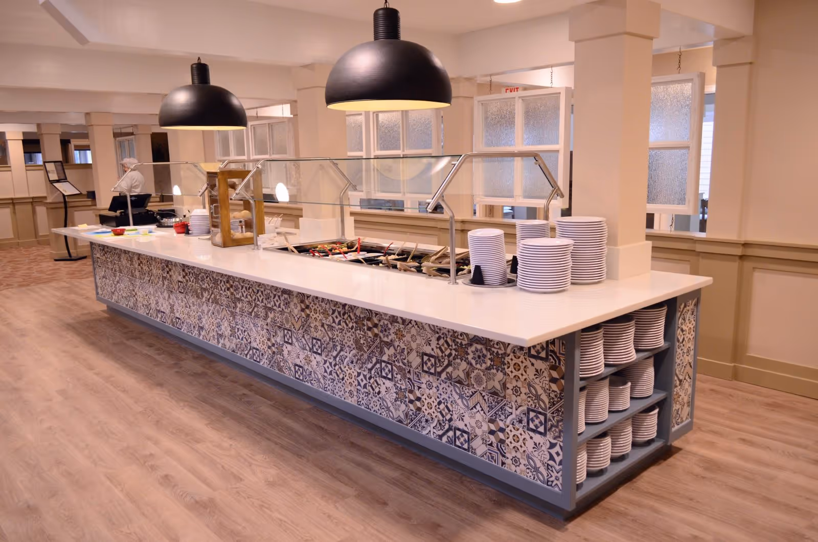 A buffet counter in a dining area with stacks of white plates on both ends. The counter has a decorative tile front and a glass sneeze guard above the food trays. Two large black pendant lights hang from the ceiling above the counter. The floor is wooden, and there is a person in the background near a wall.