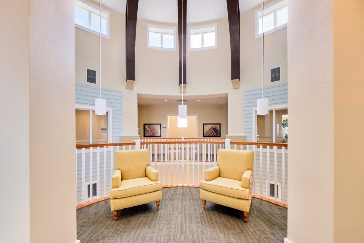 A bright and airy seating area with two yellow armchairs positioned on a carpeted floor. The space features a curved white railing, high ceilings with dark wooden beams, and several small windows near the ceiling allowing natural light to enter. Pendant lights hang from the ceiling, and framed artwork is visible on the far wall.