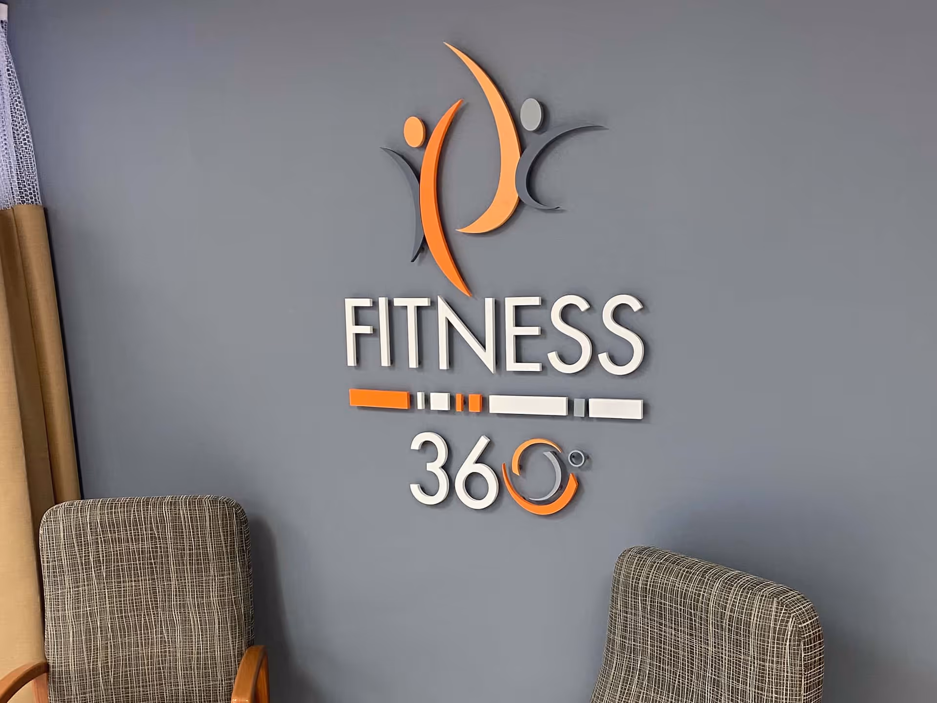 Gray wall with a 'FITNESS 360°' sign and logo above two upholstered chairs.