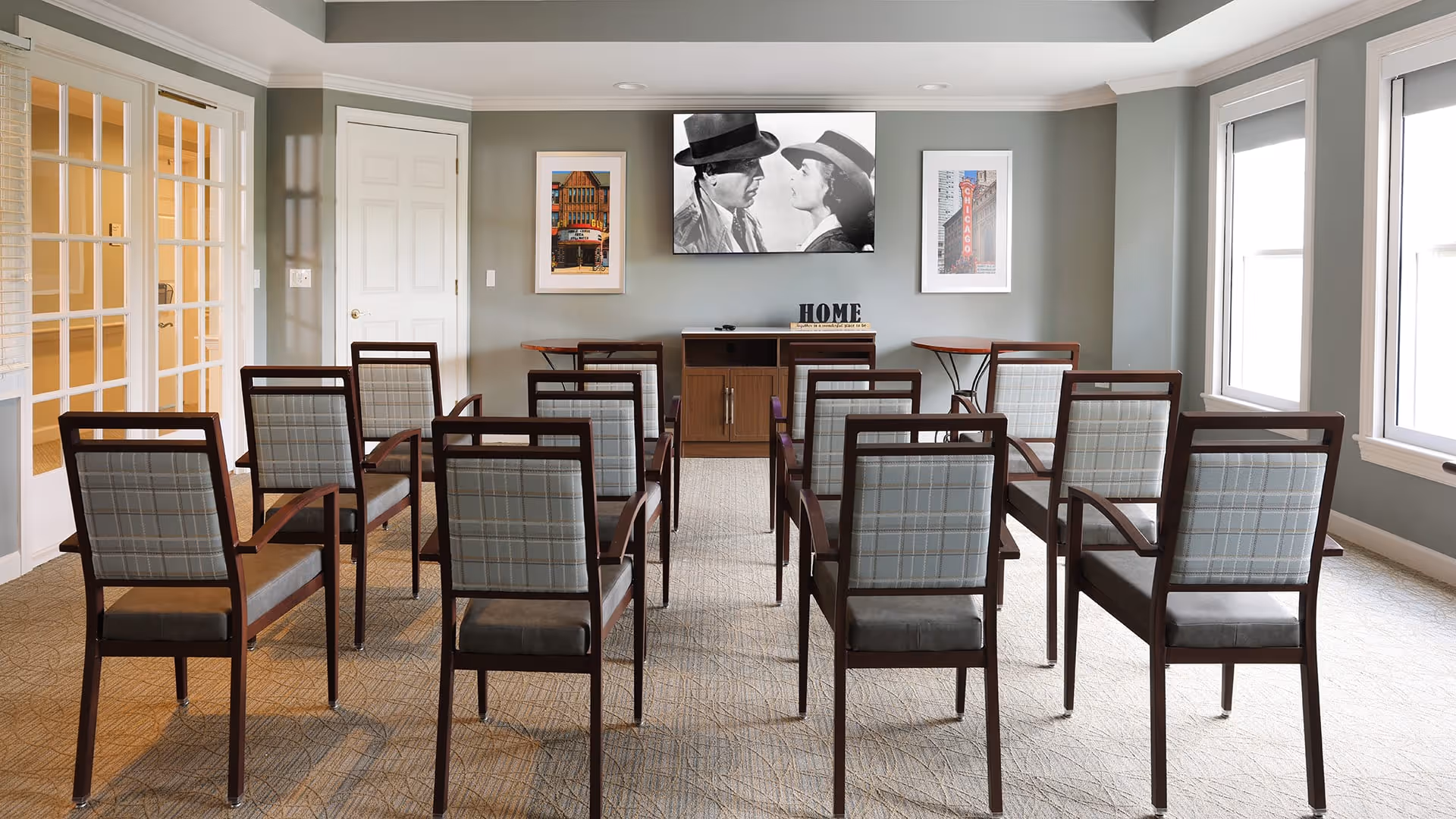 A small meeting or media room with three rows of wooden chairs facing a wall-mounted TV screen displaying a black and white movie scene. The room has light gray walls, two framed pictures on either side of the TV, a wooden cabinet below the TV with the word 'HOME' on it, and large windows on the right letting in natural light.