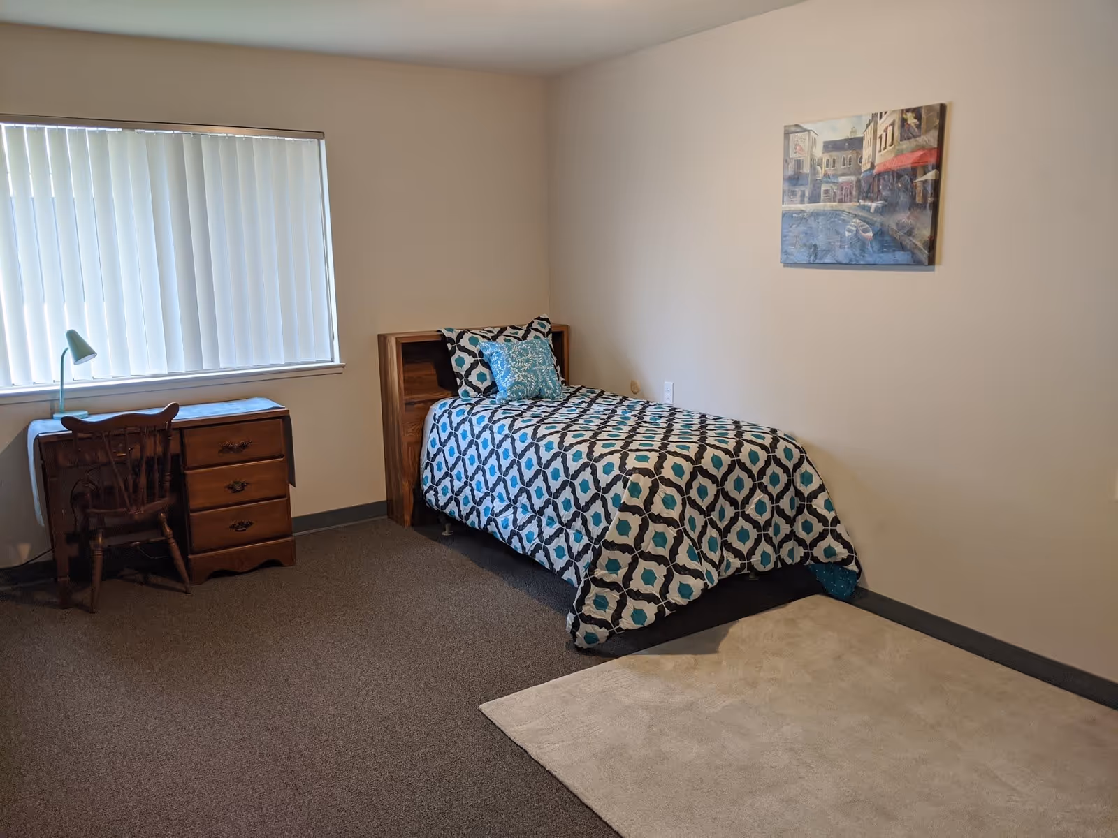 A simple bedroom with a single bed covered in a patterned blue, black, and white comforter. There is a wooden desk with a chair and a small lamp next to a window with vertical blinds. A painting depicting a street scene hangs on the beige wall above the bed. The floor is carpeted with a small beige rug near the bed.