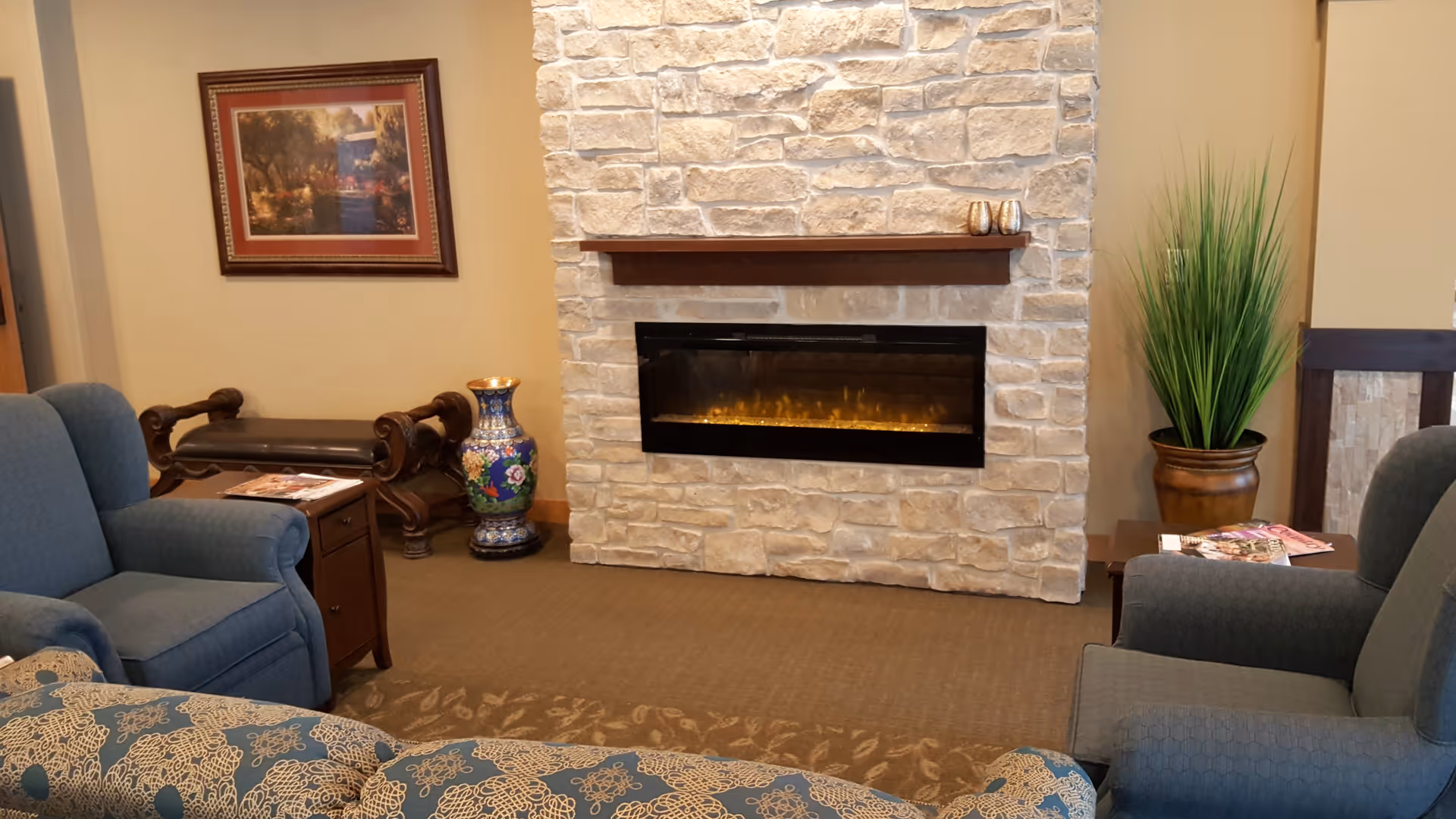 Cozy living room area with a stone fireplace in the center, flanked by two blue upholstered armchairs and a patterned sofa. A wooden side table with magazines and a tall potted plant are on the right side. On the left side, there is a decorative vase and a framed painting hanging on the beige wall.