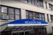 Entrance of Meadow Park Rehabilitation & Health Care Center with a blue awning displaying the facility's name. The building exterior is beige with multiple windows, some with air conditioning units.