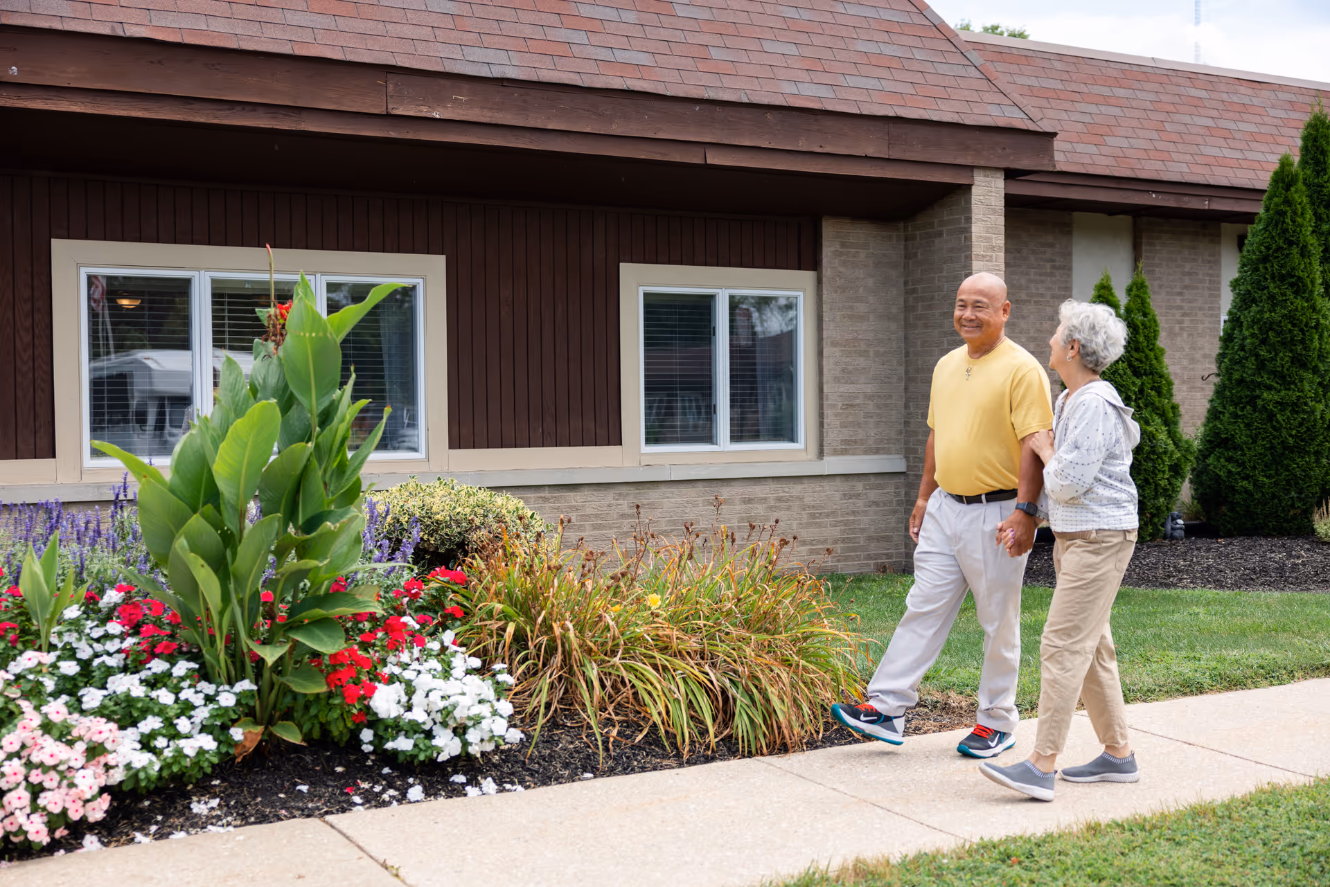 An elderly couple walking hand in hand on a sidewalk next to a garden with colorful flowers and greenery in front of a building with brown siding and windows.