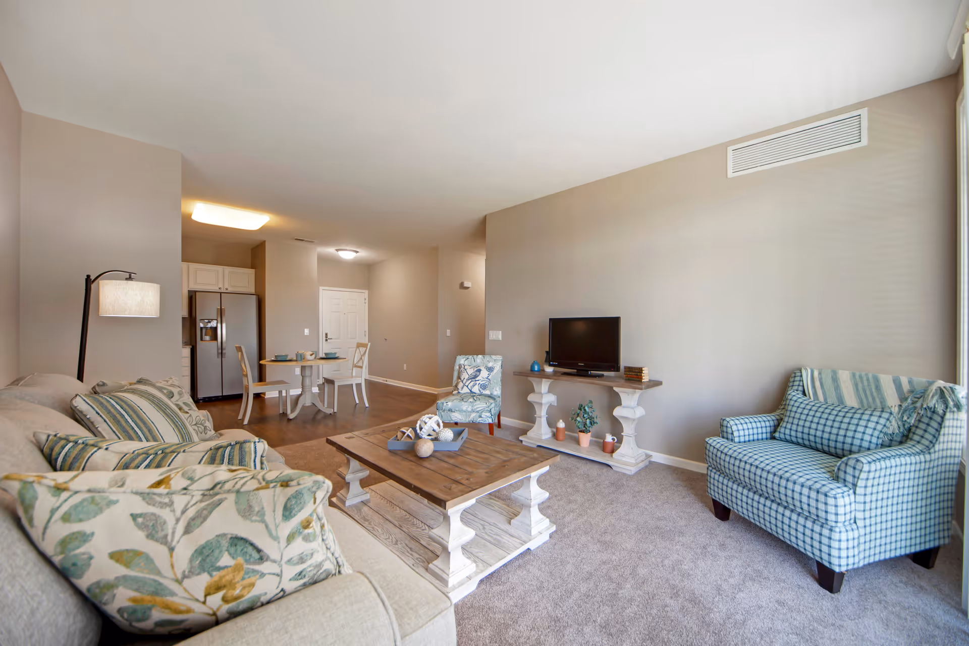 A cozy living room area with a beige sofa adorned with patterned cushions, a wooden coffee table with decorative items, a blue checkered armchair with a striped throw, and a floral patterned chair. A TV sits on a console table against a beige wall. In the background, there is a small dining area with a round table and two chairs, and a kitchen with a refrigerator. The room has carpeted flooring and neutral-colored walls.