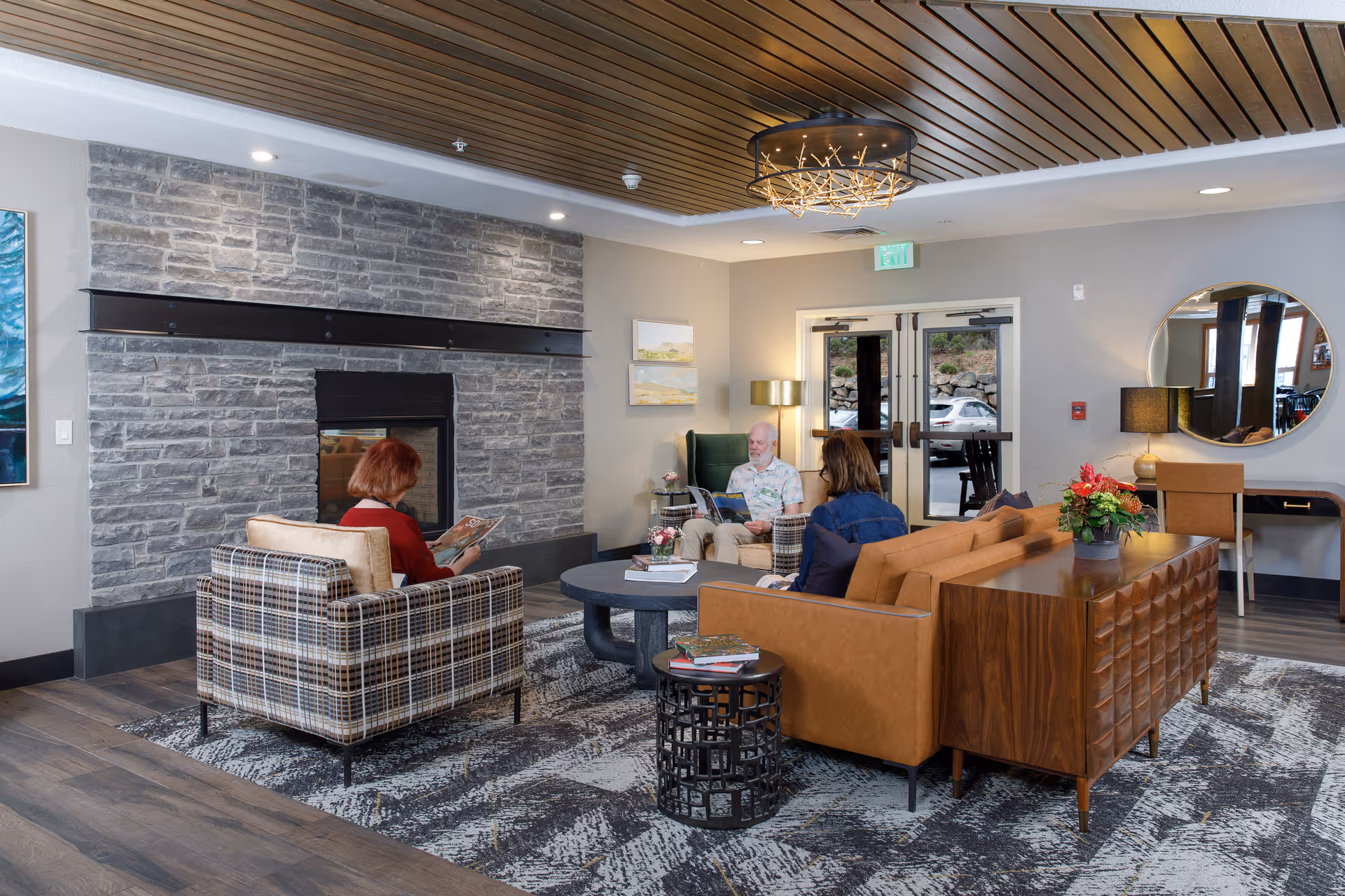 A cozy living room area in a senior living facility with three people seated and reading. The room features a stone fireplace, a plaid armchair, a brown leather sofa, a round coffee table, and a wooden sideboard with a flower arrangement. The ceiling has wooden slats and a modern chandelier. There is a large round mirror on the wall and double glass doors leading outside.