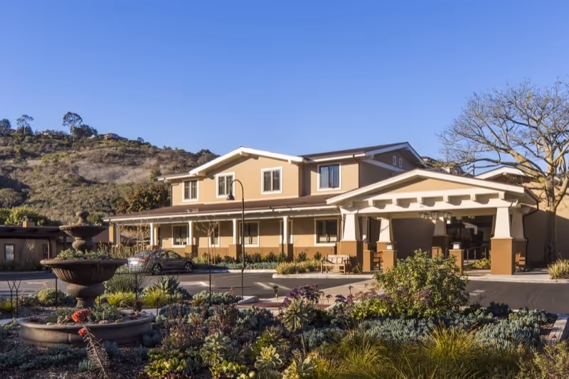 Exterior view of a two-story senior living facility building with beige walls and white trim, surrounded by landscaped gardens with various plants and a multi-tiered fountain in the foreground. Hills and clear blue sky are visible in the background.