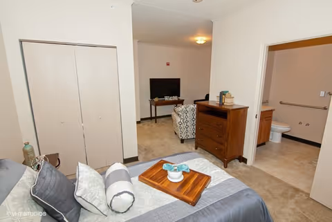Interior view of a senior living facility room at The Woodlands of Middletown showing a bed with pillows and a wooden tray, a dresser, an armchair facing a TV on a stand, and an open door leading to a bathroom with a toilet and grab bars.