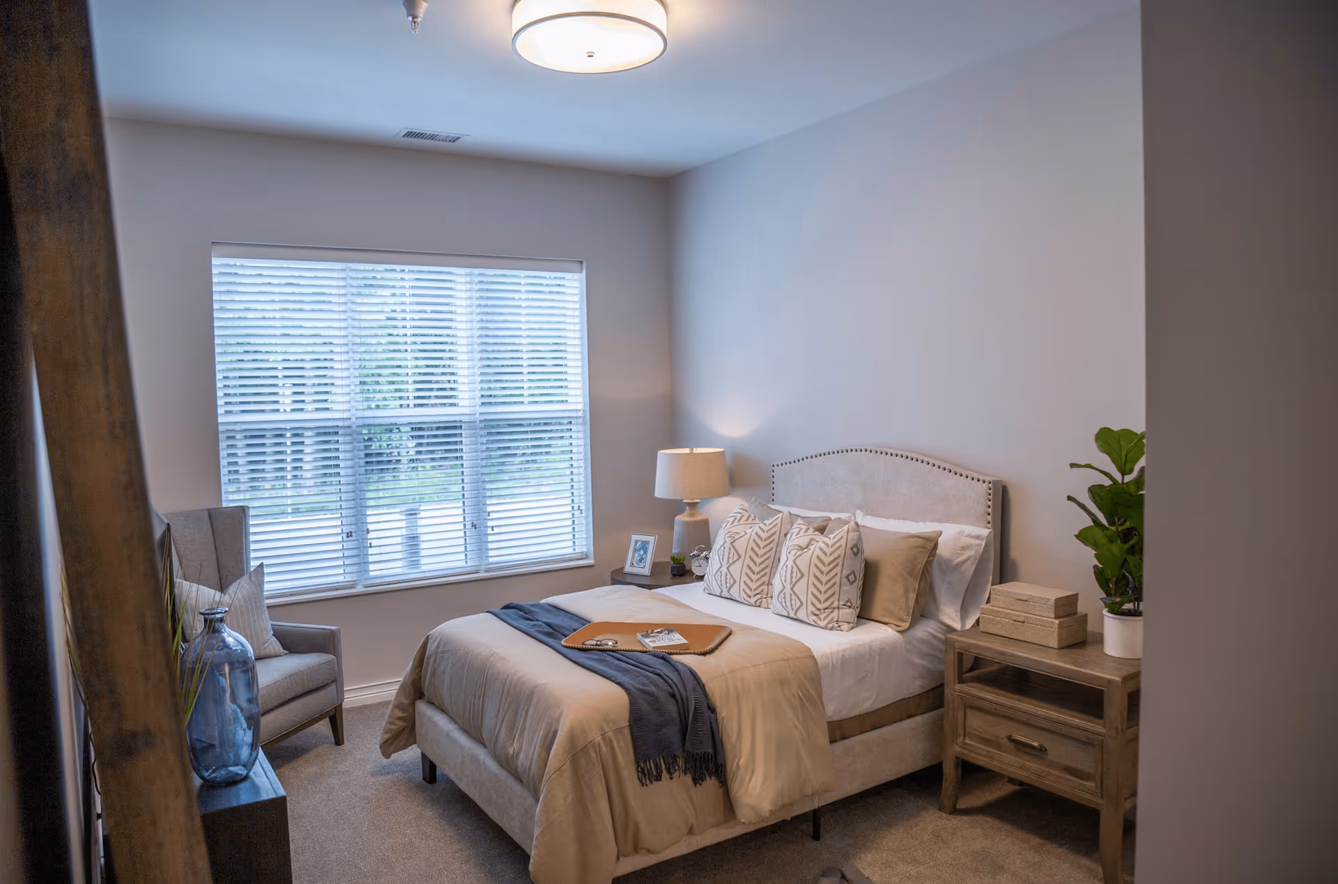 A cozy bedroom with a beige upholstered bed adorned with multiple patterned pillows and a beige comforter. A blue throw blanket and a tray with a book and glasses rest on the bed. To the right of the bed is a wooden nightstand with a lamp, two decorative boxes, and a potted plant. A large window with white blinds lets in natural light. A gray armchair with a pillow and a large blue vase are positioned near the window. The room has neutral-colored walls and carpeted flooring.