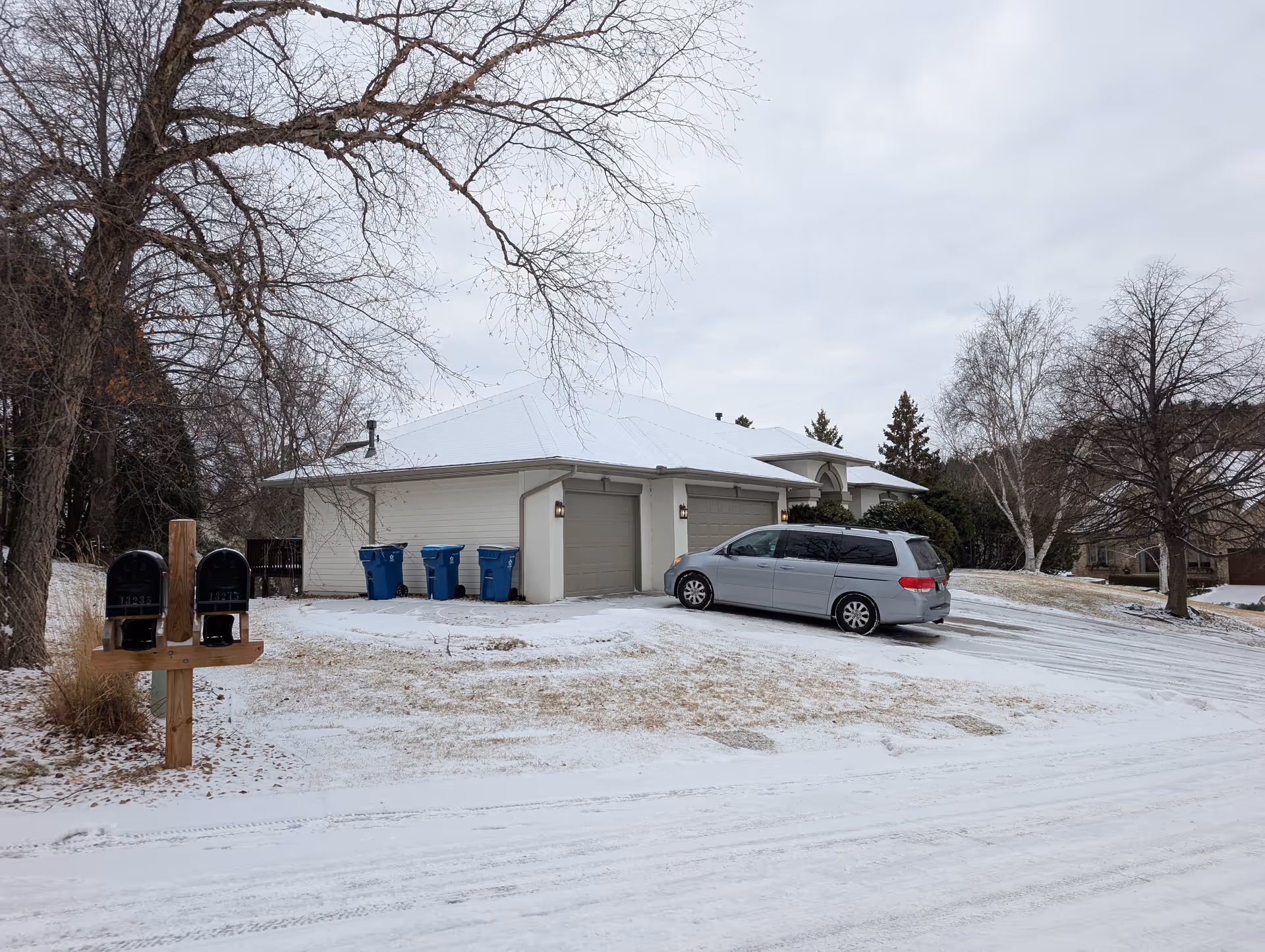 A snowy residential driveway with a gray minivan parked in front of a two-car garage attached to a light-colored house. There are three blue recycling bins lined up near the garage. Two black mailboxes mounted on a wooden post are visible on the left side of the image. Leafless trees and a cloudy sky are in the background.