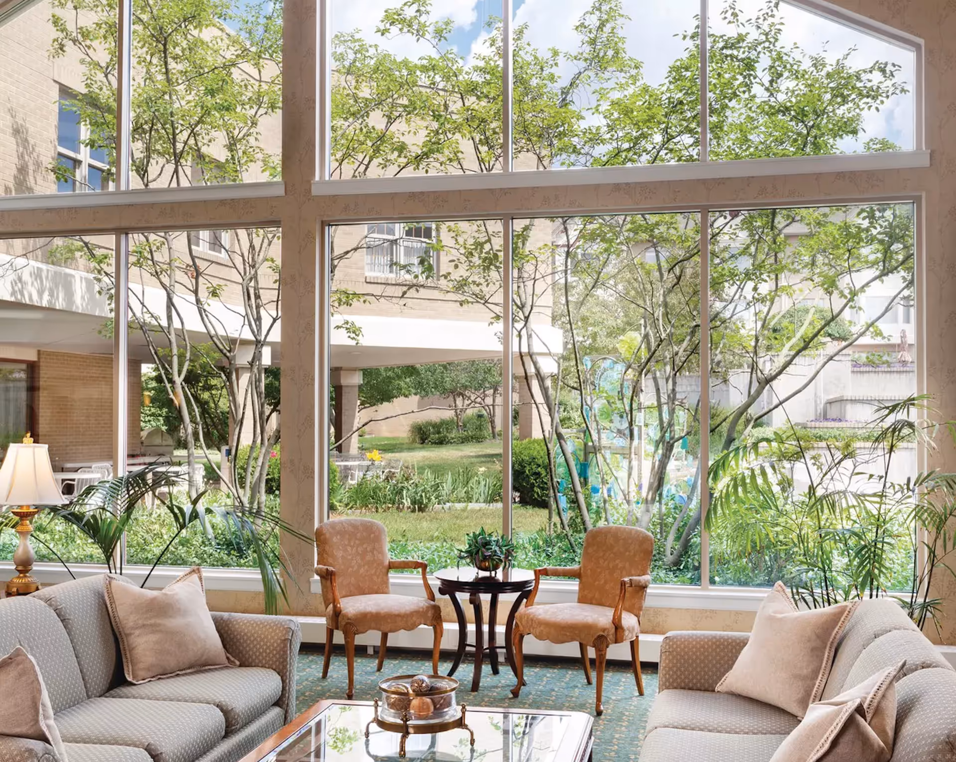 A bright and cozy living room area with two beige sofas, two upholstered armchairs, a glass coffee table with decorative items, and large floor-to-ceiling windows showing a green garden outside.