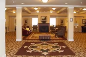 Spacious common living room with upholstered chairs and sofas arranged around a central fireplace beneath a coffered ceiling and patterned carpet.