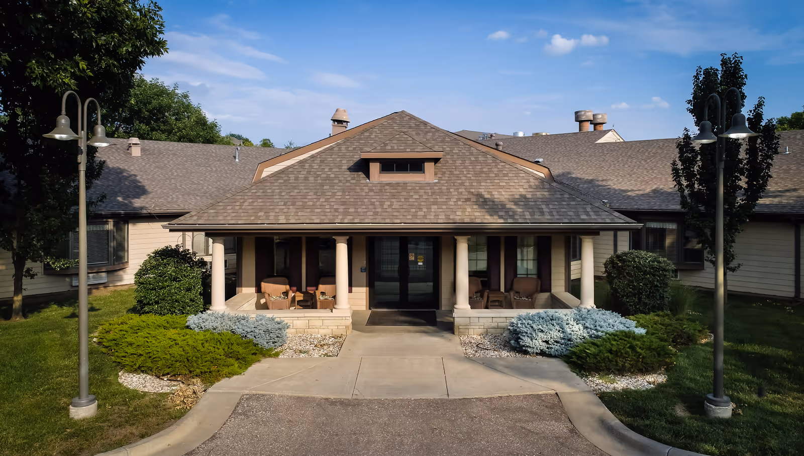 Front exterior view of a single-story assisted living facility building with a covered entrance supported by white columns. There are outdoor chairs on the porch, landscaped bushes, and two lamp posts on either side of the walkway leading to the entrance under a blue sky with scattered clouds.