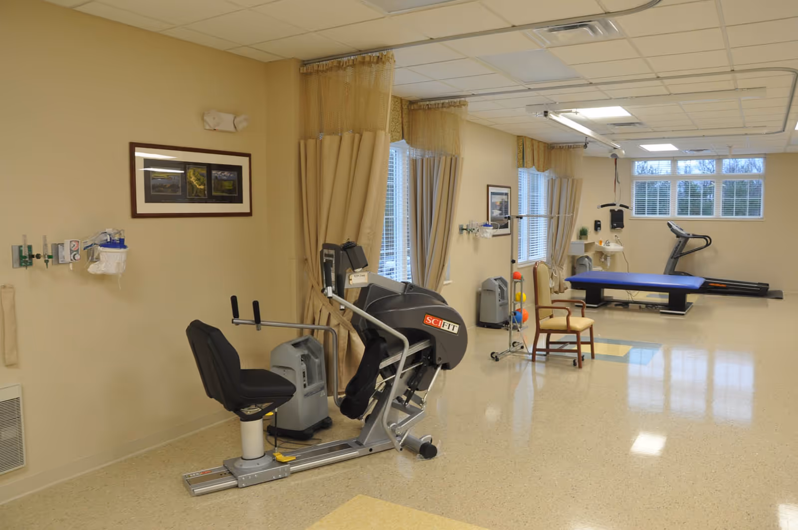A rehabilitation room with exercise equipment including a seated leg press machine, a treadmill, and a therapy table. The room has beige walls, large windows with curtains, a chair, and medical supplies mounted on the wall.