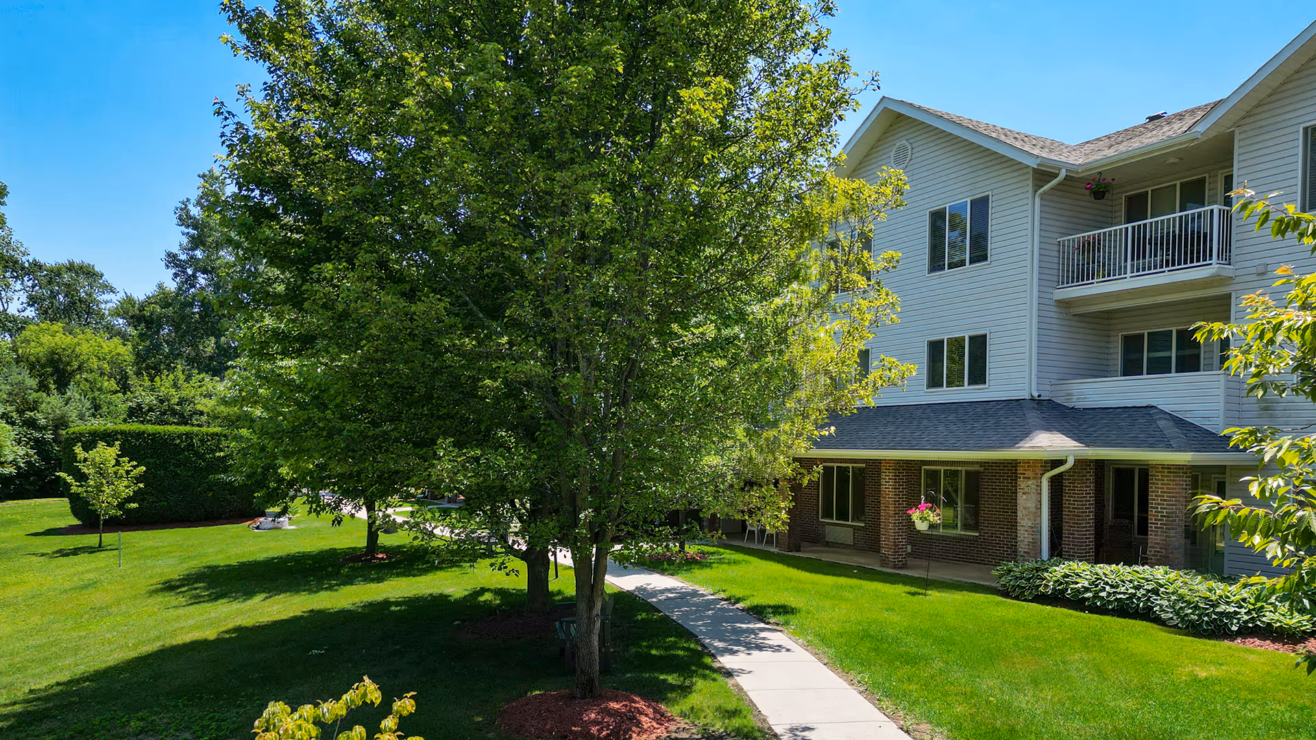 A sunny outdoor view of Holiday Blue Water Lodge showing a well-maintained green lawn with trees and shrubs, a concrete walkway leading to the building entrance, and a multi-story residential building with balconies and hanging flower pots.