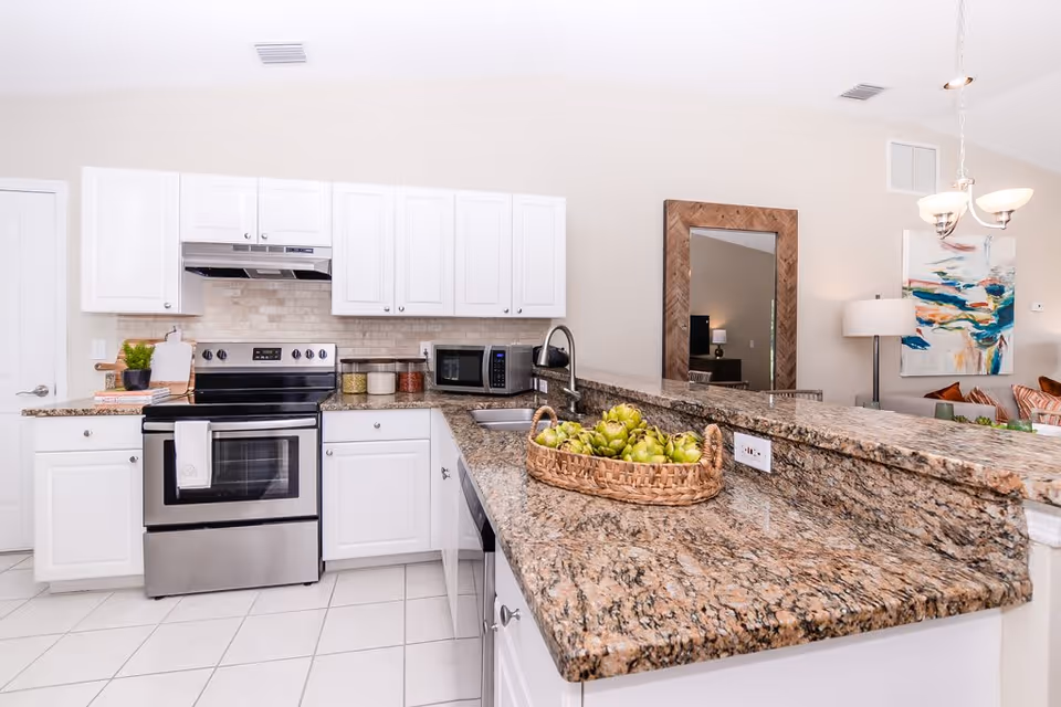 Bright kitchen with white cabinets, stainless steel stove and microwave, granite countertops, a basket of green artichokes on the counter, and a view into a living area with a large mirror, floor lamp, and colorful abstract painting.