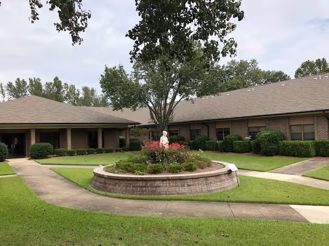 A courtyard area of a nursing center with a circular raised brick planter in the center containing a tree and flowers. Surrounding the planter is a concrete walkway and well-maintained grass. The building in the background is a single-story brick structure with multiple windows and a sloped roof.