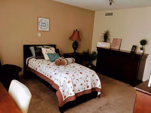 A cozy bedroom with a single bed covered in a white quilt with autumn leaf patterns. The bed has multiple pillows including a cylindrical bolster pillow. Next to the bed is a dark wooden nightstand with a decorative lamp and some plants. On the wall above the bed is a framed sign that reads 'Simply Blessed'. Across from the bed is a dark wooden dresser with decorative items including a 'FAMILY' sign, a framed quote, and a small potted plant. The room has beige walls and carpeted floor.