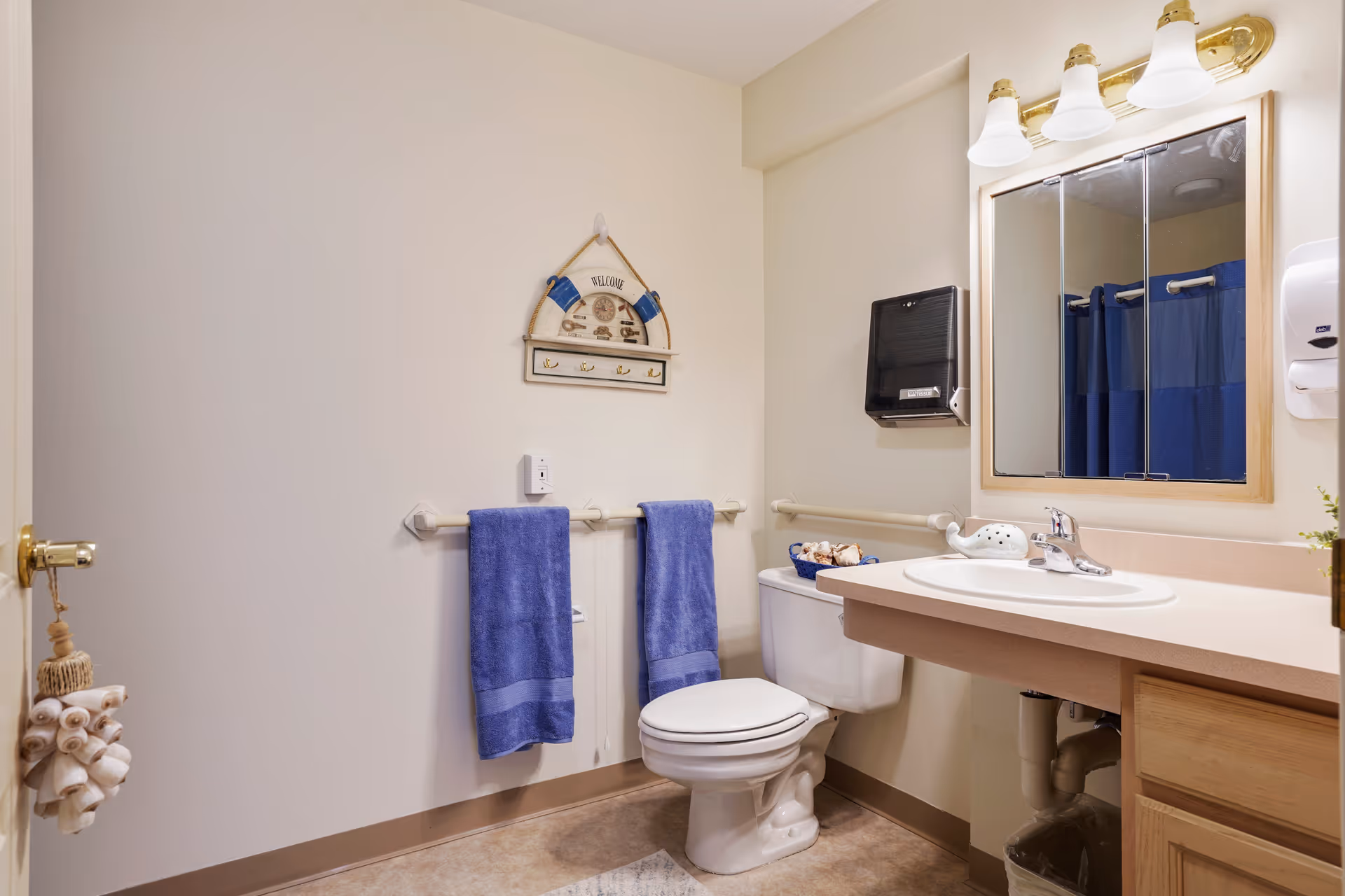 A clean bathroom with beige walls and floor. There is a white toilet with a blue basket on top, a sink with a beige countertop, a mirror with three lights above it, and two blue towels hanging on a towel rack. A decorative wall hanging with the word 'Welcome' is above the towels. A soap dispenser and paper towel dispenser are mounted on the wall.