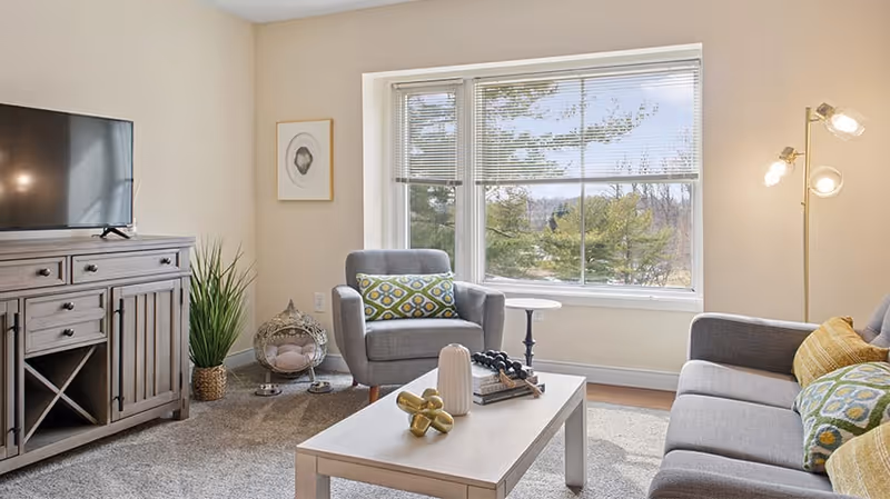 A cozy living room with a large window showing trees outside. The room features a gray armchair with a patterned cushion, a gray sofa with yellow and patterned cushions, a wooden coffee table with decorative items, a wooden TV stand with a flat-screen TV, a tall floor lamp with three lights, a potted plant, and a small side table.