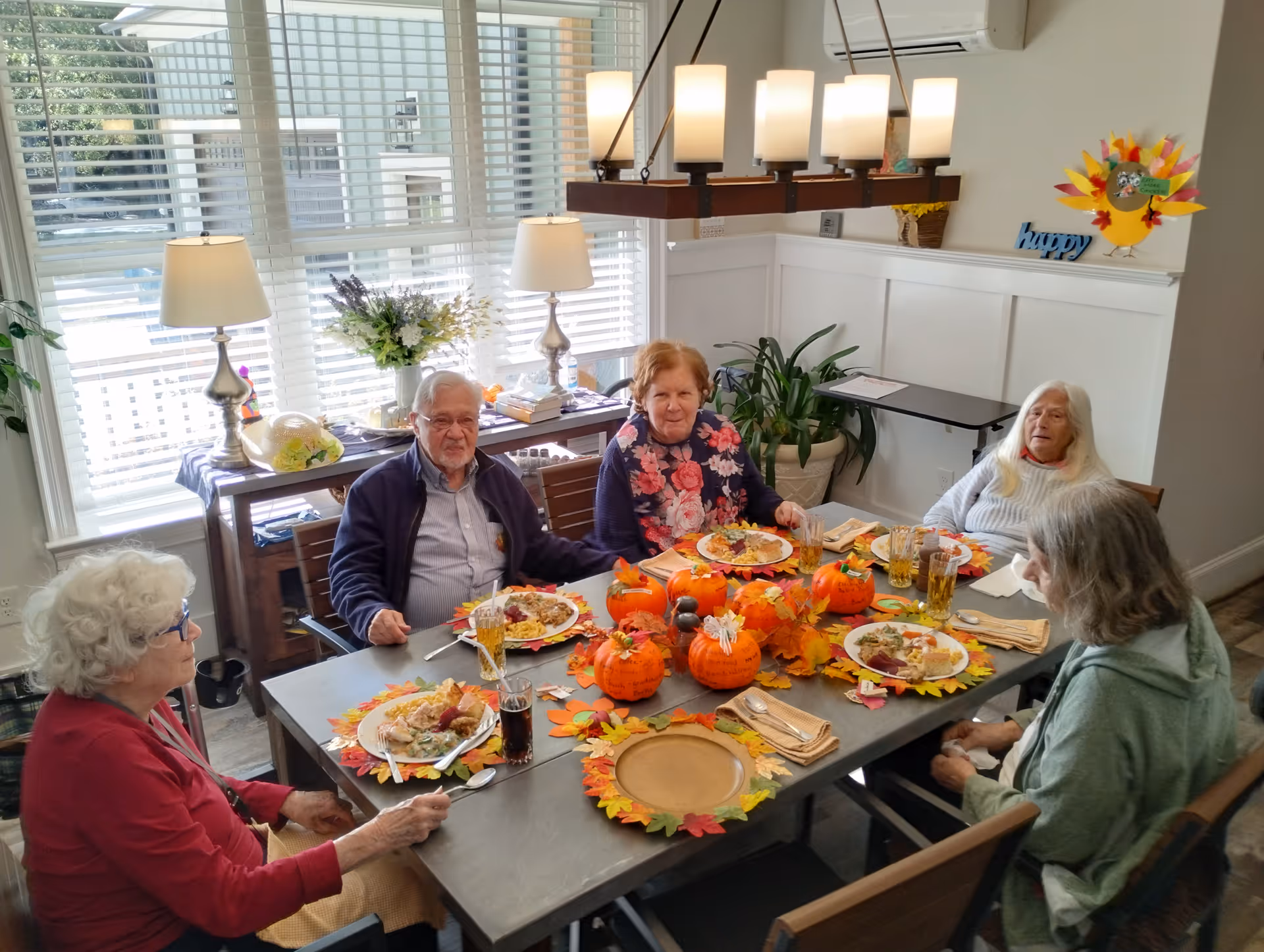 Five elderly residents sit around a fall-decorated dining table enjoying a meal in a bright dining room.