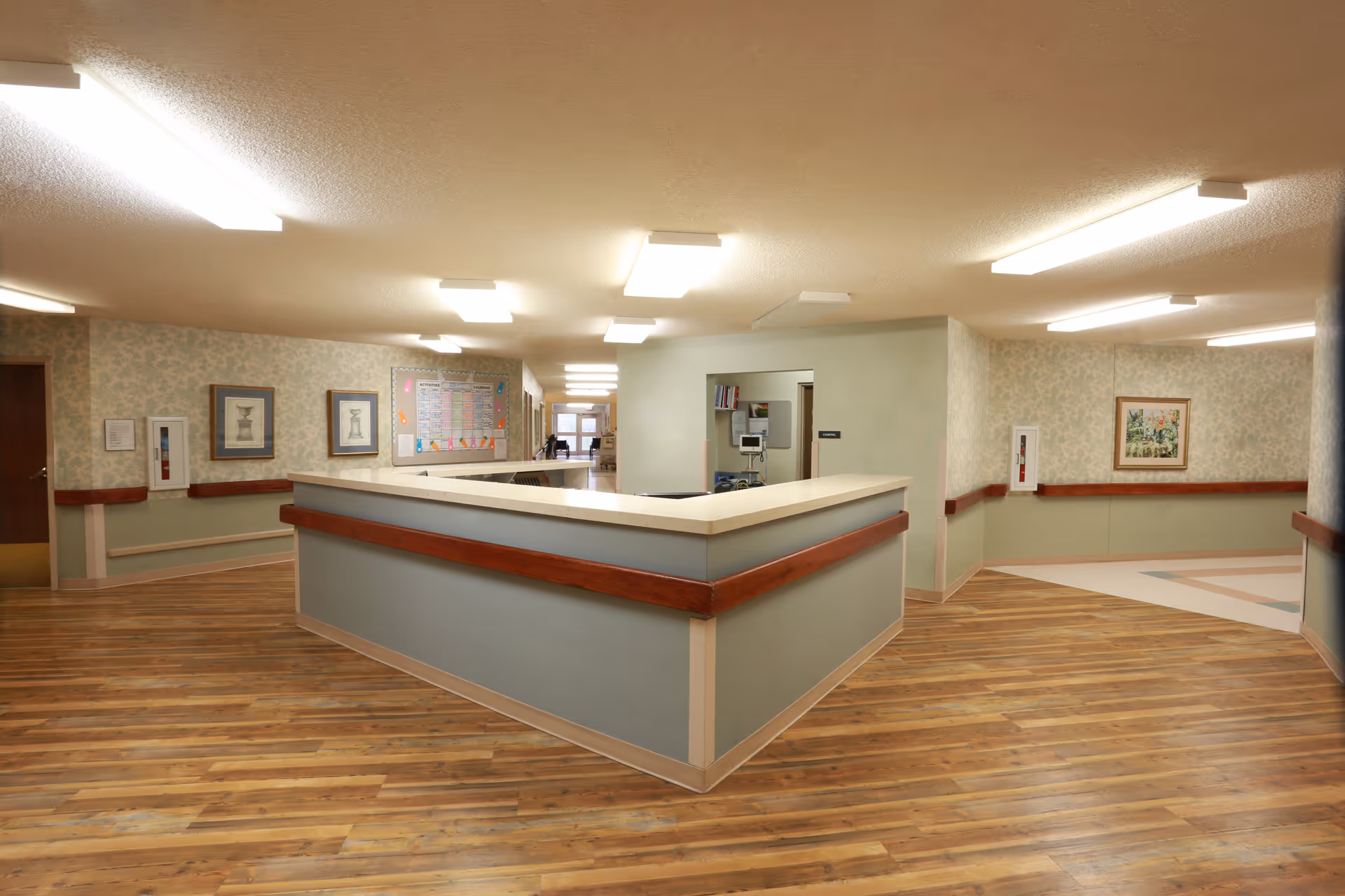 Interior view of a senior living facility hallway with a central nurse's station featuring a light blue counter with wooden trim. The floor is wood-patterned, and the walls are decorated with framed artwork and a bulletin board. Fluorescent ceiling lights illuminate the space.