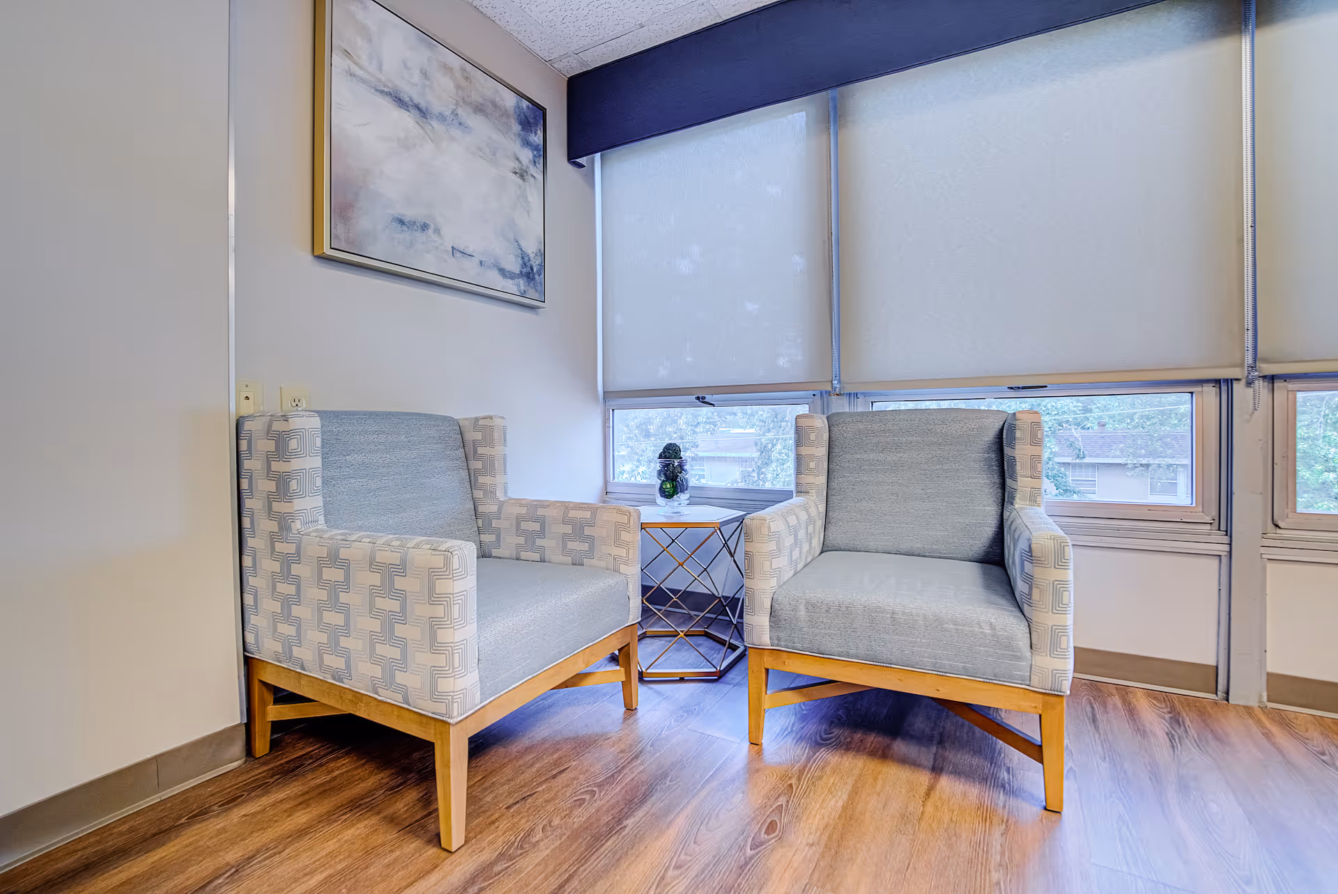 Two patterned armchairs with wooden legs are placed on a wooden floor near a window with closed blinds. A small round side table with a decorative item is positioned between the chairs. A framed abstract painting hangs on the wall above one of the chairs.