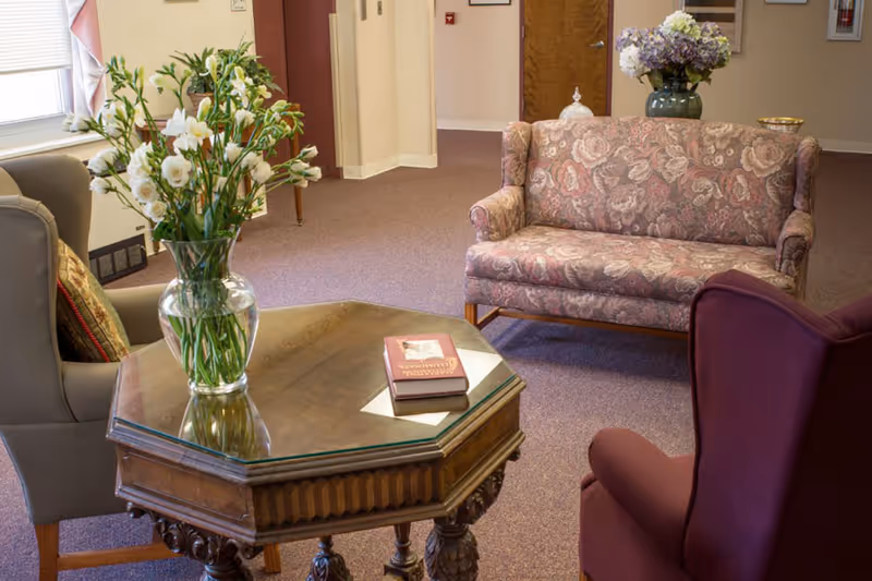A cozy seating area with upholstered chairs and a floral loveseat around a wooden octagonal coffee table topped with a vase of white flowers and a book.
