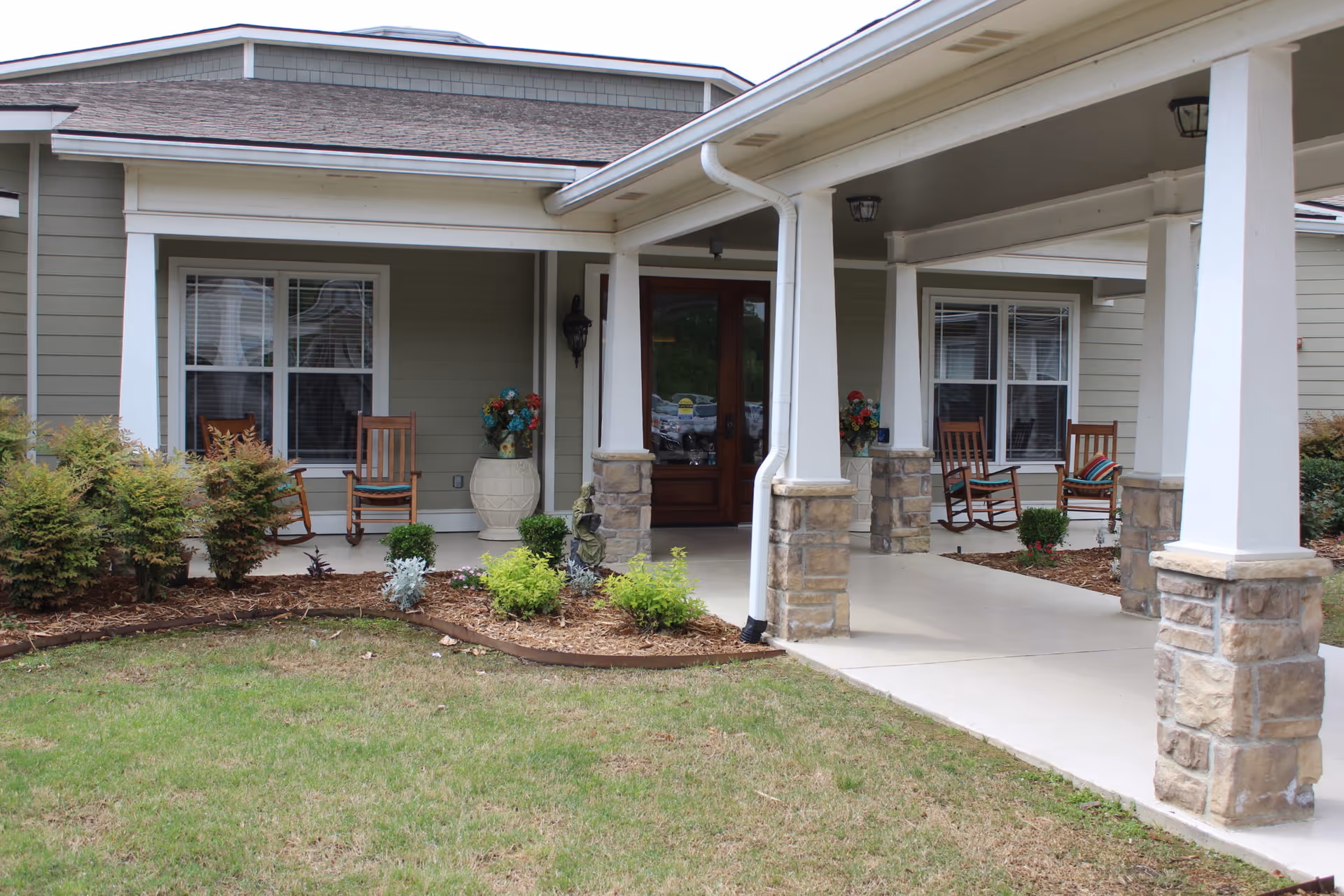Front porch area of a senior living facility with a covered walkway supported by white columns with stone bases. There are wooden rocking chairs with cushions placed near windows on either side of a wooden door. Decorative flower pots and small shrubs are planted along the edge of the porch and lawn.