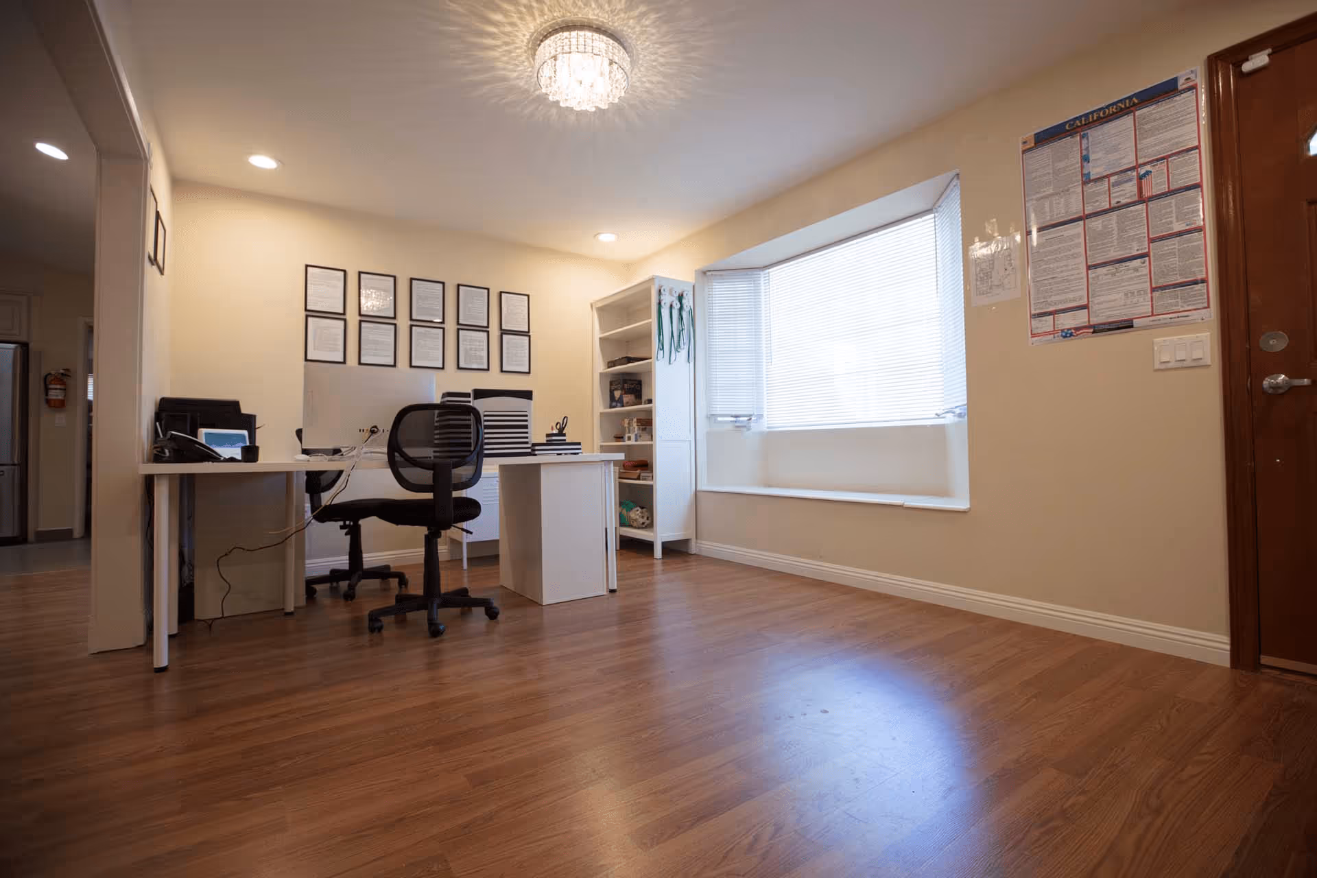 A clean, well-lit office space with wooden flooring, a white desk with a black office chair, a computer monitor, and office supplies. The room has a large window with blinds, a white shelving unit with various items, framed documents on the wall, and a wooden door with a California labor law poster beside it.