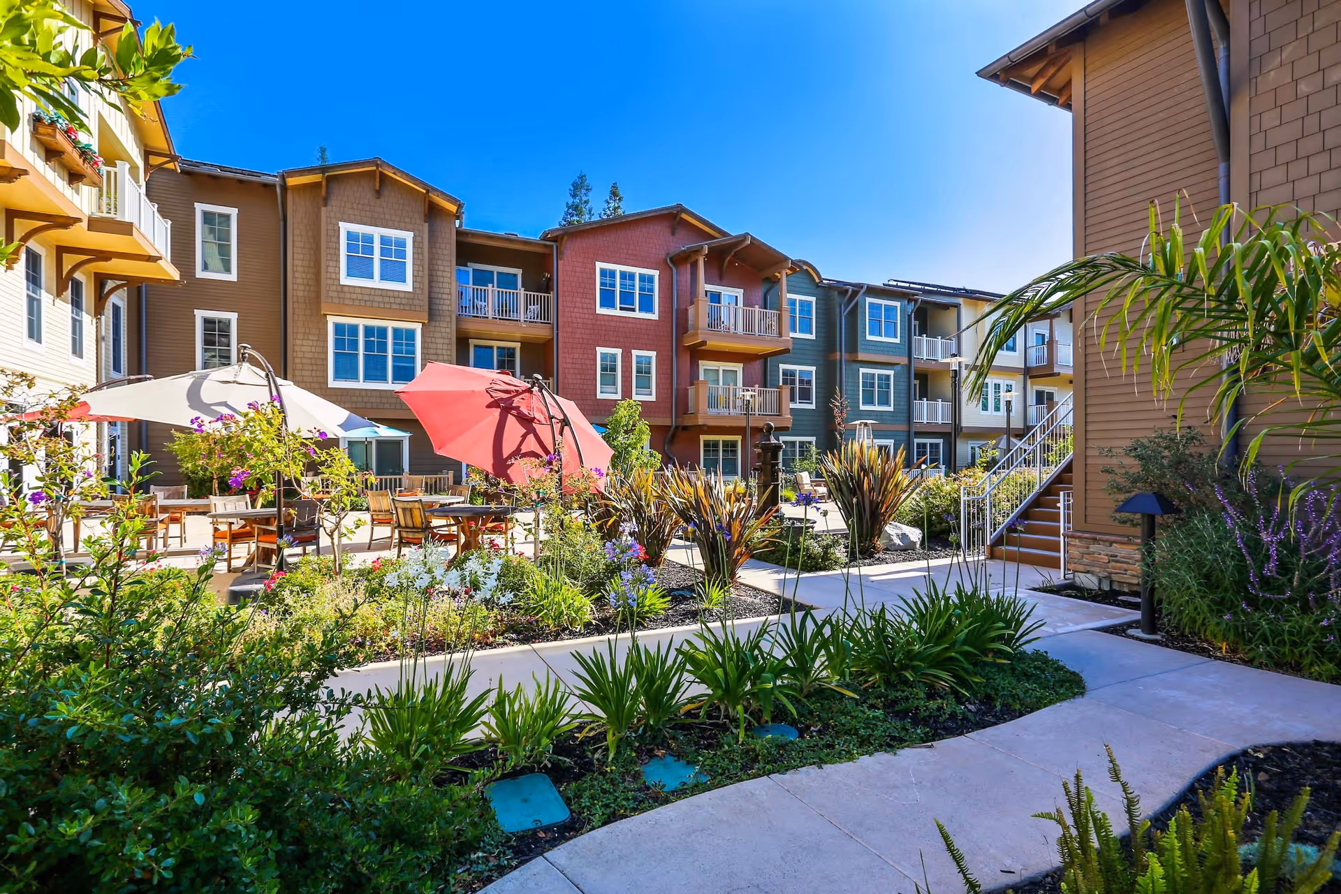 Sunny landscaped courtyard with colorful multi-story residential buildings, patio tables, umbrellas, and planted walkways.