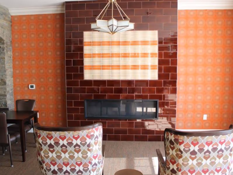 A cozy interior room with two patterned armchairs facing a modern rectangular fireplace set in a wall of glossy dark red tiles. The walls on either side are covered with orange wallpaper featuring a circular pattern. A geometric pendant light hangs above the fireplace, and a small dark wooden table with chairs is visible to the left.